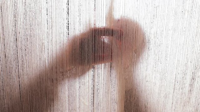 Aesthetic and abstract shot of an artisan's hands working in silhouette behind the vertical warp threads on a traditional weaving loom. Handicrafts, textiles, and labor concept.