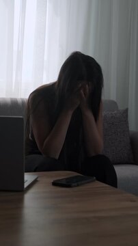 Depressed woman with head down sitting on sofa in despair at home next to laptop and phone, illustrating mental fatigue. Realistic depiction of freelance burnout, vertical
