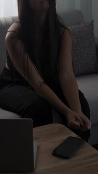 Procrastination and work avoidance concept with young woman sitting on sofa at home feeling mental resistance next to laptop and phone, vertical