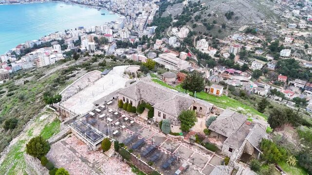Aerial tracking shot flying forward over the stone round tower and walls of Lekuresi Castle, revealing the coastal city of Sarande and the Ionian Sea in Albania