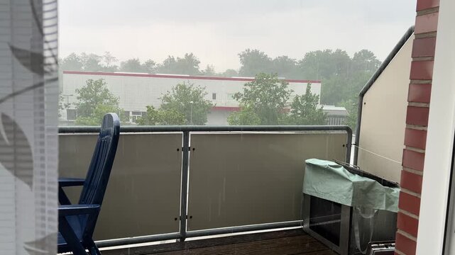 View from a window of heavy rain falling on an apartment balcony with a blue plastic chair, looking out towards green trees and a building under a overcast sky.