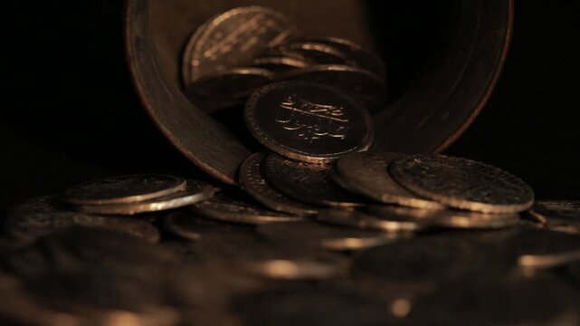 Dramatic, low-light close-up of old, authentic coins with Arabic/Ottoman inscriptions spilling out of a metal container. Treasure, history, and ancient era concept.