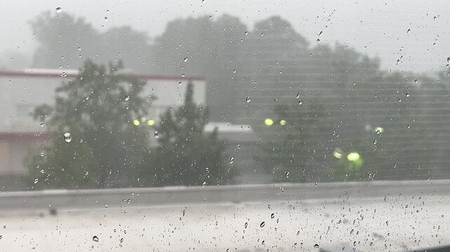 View from a window of heavy rain falling on an apartment balcony with a blue plastic chair, looking out towards green trees and a building under a overcast sky.
