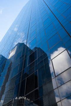 A blue glass skyscraper facade holds reflection of clouds and a neighboring tower, with windows climbing into the sky above the city edge