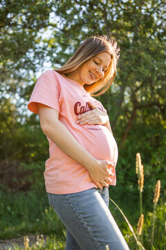 Expectant woman smiling outdoors, touching baby bump in casual maternity portrait during golden hour