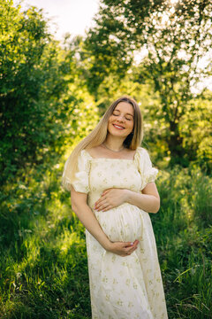Happy pregnant woman in floral maternity dress standing in sunlit green meadow, smiling peacefully during summer
