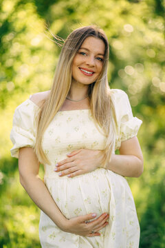Pregnant woman smiling outdoors holding baby bump in floral maternity dress during summer light
