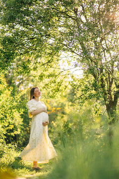 Pregnant woman in a flowing white dress enjoying sunlight in a green nature maternity portrait