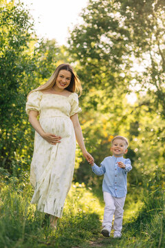 Pregnant mother walking hand in hand with happy young son in sunlit summer meadow