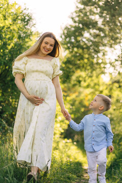 Pregnant mother walking hand in hand with son in sunny green park enjoying maternity moments