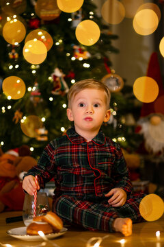 Little boy in plaid pajamas sitting by a Christmas tree with lights and treats
