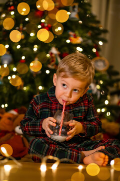 Young boy in plaid pajamas sipping milk by a decorated Christmas tree with warm holiday lights