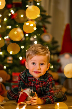 Little boy in plaid pajamas smiling by a decorated Christmas tree with warm bokeh lights