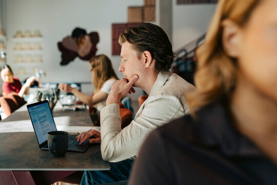 Businessman with hand on chin working on laptop while sitting at desk in shared working space