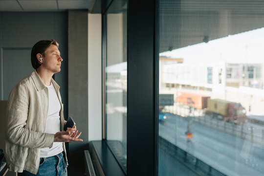 Young businessman wearing wireless earbuds and looking through glass window at office