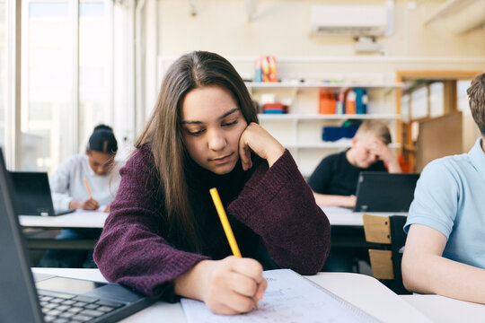 Teenage girl writing on book while leaning on elbow at desk in classroom