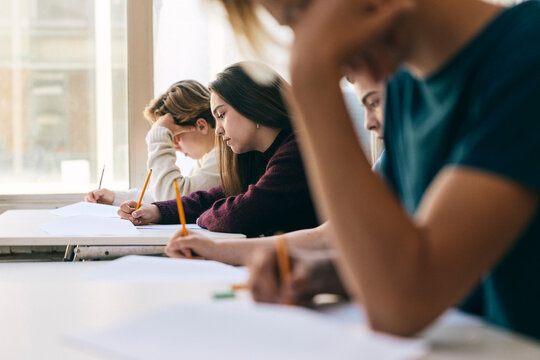 Focused teenage schoolgirl writing exam paper with friends while sitting at desk in classroom