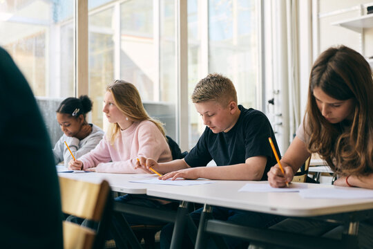 Focused schoolboy writing paper while sitting at desk in exam hall