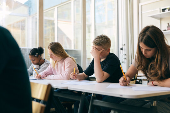 Stressed schoolboy writing exam paper while sitting with classmates at desk