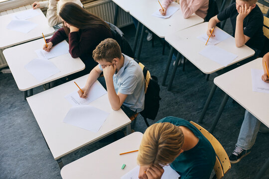 High angle view of teenage junior high students writing exam paper while sitting at desk in classroom