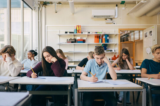 Multiracial male and female junior high students writing education exam while sitting in classroom