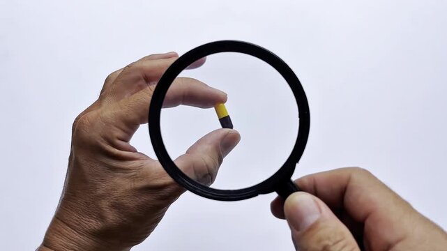 Close-up of a hand holding a capsule under a magnifying glass, symbolizing medicine inspection, healthcare research, pharmaceutical quality control, and wellness.