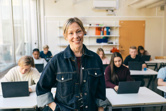 Portrait of smiling female teacher standing with hand in pocket in classroom at junior high school
