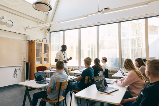 Male professor teaching students sitting with laptop in classroom at junior high school