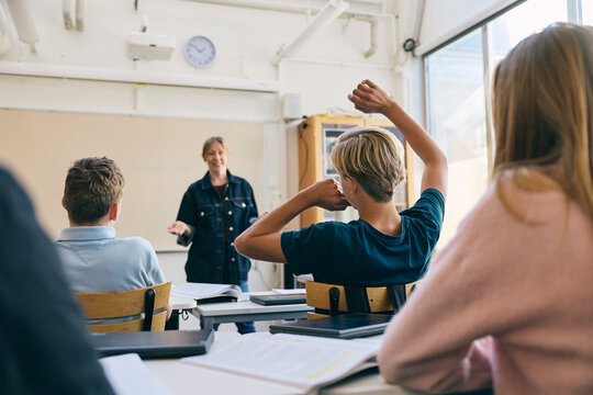 Rear view of teenage boy raising hand while asking question to female teacher in classroom at school