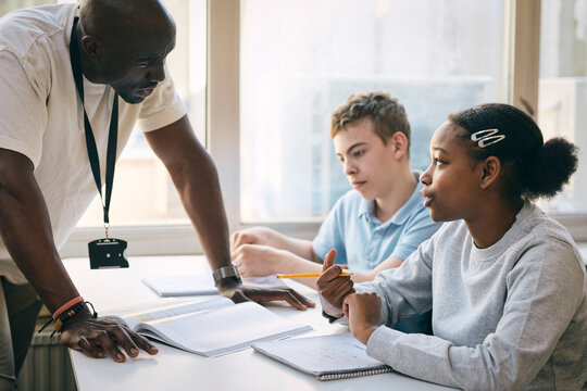 Male teacher listening to teenage girl asking question while studying in classroom at school