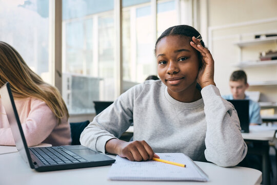 Portrait of smiling teenage girl sitting with laptop and book in classroom at junior high school