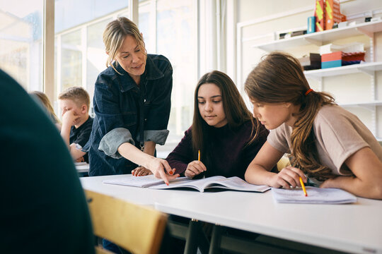 Female teacher assisting teenage girls while pointing at book on desk in classroom