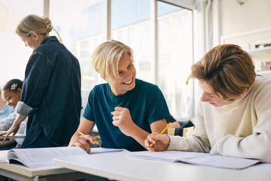 Happy teenage boy celebrating after solving math equation while sitting with friend at desk in school