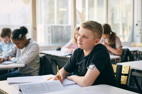 Thoughtful male student wearing glucose monitor on arm sitting at desk while studying in school