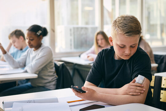 Junior high school student checking blood sugar level through glucose meter while sitting at desk with books in classroo