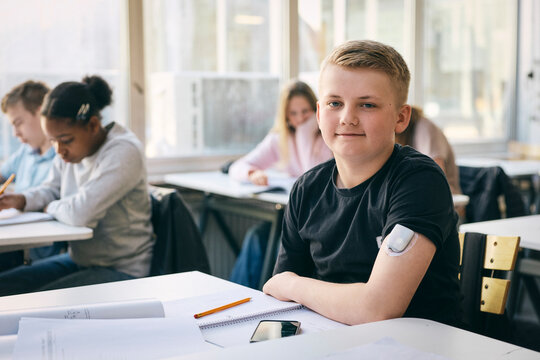 Portrait of smiling teenage boy having chronic illness wearing glucose monitor on arm while sitting at desk in classroom
