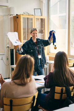 Smiling female teacher showing certificates and medals in class at school