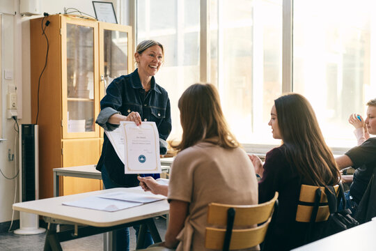 Happy female teacher showing certificate to teenage girl sitting at desk in classroom at junior high school