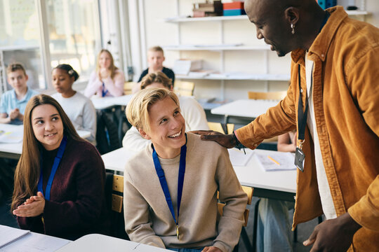 Male teacher talking with smiling teenage boy medalist sitting at desk in junior high school