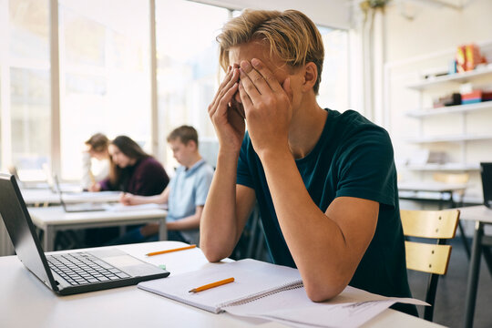 Tired teenage boy sitting with hands covered face at desk in classroom