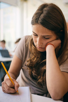 Teenage girl solving math equation in book while leaning on elbow at classroom desk