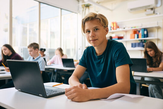 Portrait of smiling male student sitting at desk with laptop in classroom