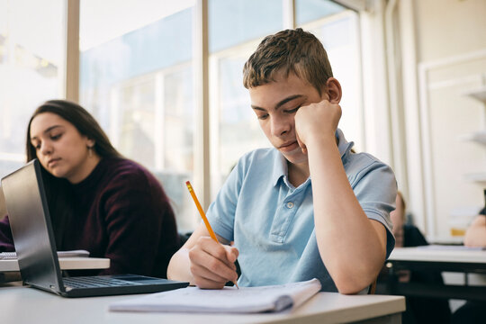 Teenage boy writing on book while sitting at desk with laptop in school classroom