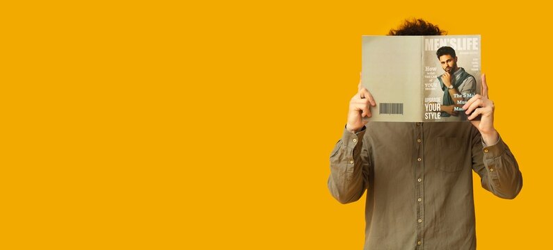 Young man covering face with lifestyle magazine while standing against bright orange background, anonymous reader holding open publication, fashion and media concept with copy space