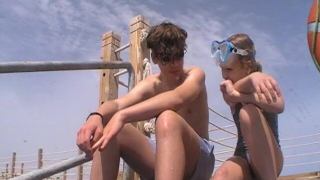 A teenager and a young girl sit side by side on a wooden sea pier with diving masks on their heads resting in the summer sun filmed on vintage analog video with retro tones