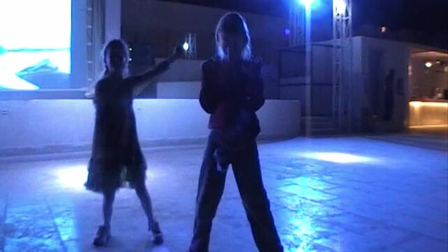 Two children stand as dark silhouettes on a glowing blue dance floor during an evening resort disco with stage lights behind, captured on vintage analog video with retro grain