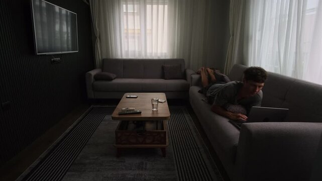 Young man spending time on laptop in small apartment changing positions on couch relaxed home screen time casual digital lifestyle and everyday indoor leisure