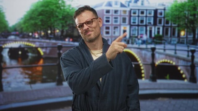 Man in a dark robe points right index finger upward on a street by a lit canal bridge and row houses, wearing glasses and smirking; amused curiosity.