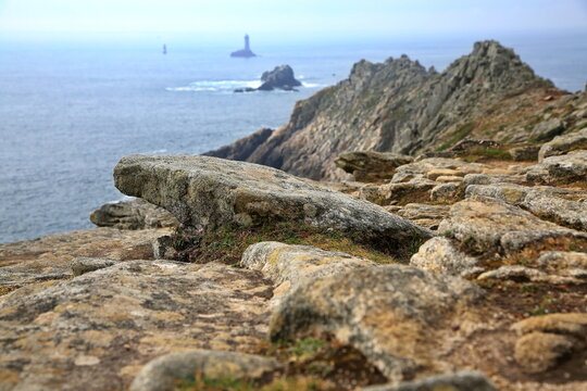 Raue Felsklippen fallen steil in den Atlantik ab, w&auml;hrend im Hintergrund die ber&uuml;hmten Leuchtt&uuml;rme Phare de la Vieille und T&eacute;vennec aus den unruhigen Wellen ragen.