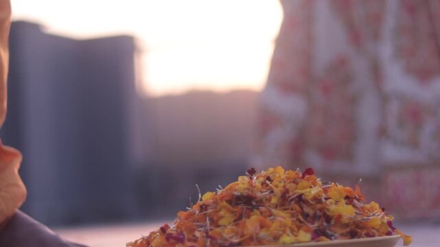 Plate filled with colorful flower petals in focus while young Indian couple in Radha Krishna attire run joyfully on rooftop terrace in background